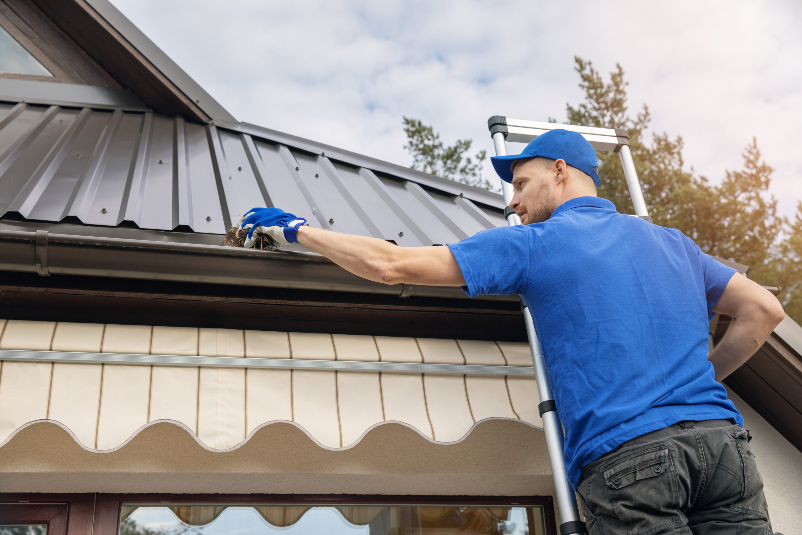 Worker on ladder cleaning gutters on metal roof — risky for gutters and personal safety